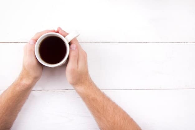 An overhead shot of a person's hands holding a cup of tea or coffee while writing in a journal. The journaling is done on a wooden table with cozy lighting and other decorative elements such as reading glasses and a pen.