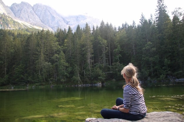 A person sitting peacefully by a lake, meditating with their eyes closed, surrounded by lush greenery and mountains in the background.