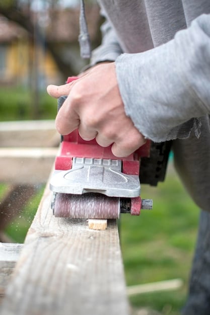 A close-up shot of a person sanding down a pallet with an electric sander. The focus is on the texture of the wood and the dust being removed, highlighting the preparation phase of the DIY project. Safety glasses and gloves are worn, emphasizing the importance of safety during the sanding process.