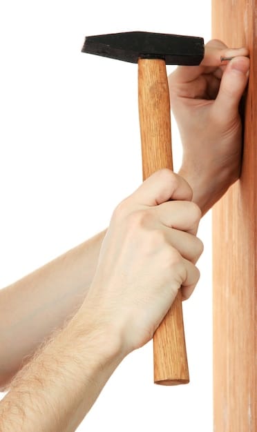 A close-up shot of a person's hands screwing together a wooden frame for a raised garden bed. The wood is rough, likely reclaimed pallet wood, and the focus is on the act of construction. The background shows a partially completed garden bed and various tools.