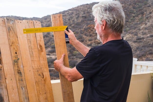 A person measuring the length and height of a backyard fence with a measuring tape, with a notepad and pen nearby to record the measurements. The background shows a garden with plants and outdoor furniture.