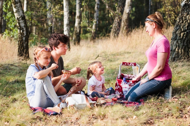 A family is having a picnic in a scenic area of a national park. They are sitting on a blanket, surrounded by trees and mountains. The picnic basket is open, revealing sandwiches, fruits, and drinks. The family looks relaxed and happy, enjoying their meal amidst nature.