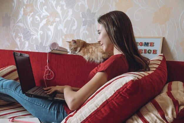 A young woman is working on her laptop on her couch, with her playful tabby cat sitting on her desk, playfully trying to get attention. The woman is smiling and petting the cat, while attending a virtual meeting.
